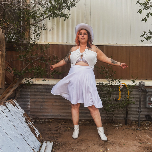 Person wearing a white dress and hat in an outdoor setting with a corrugated metal wall.