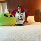 A person sitting on a bed in a bedroom, wearing a white graphic t-shirt under a red plaid flannel shirt. A large green alien plush toy is on the bed next to them.