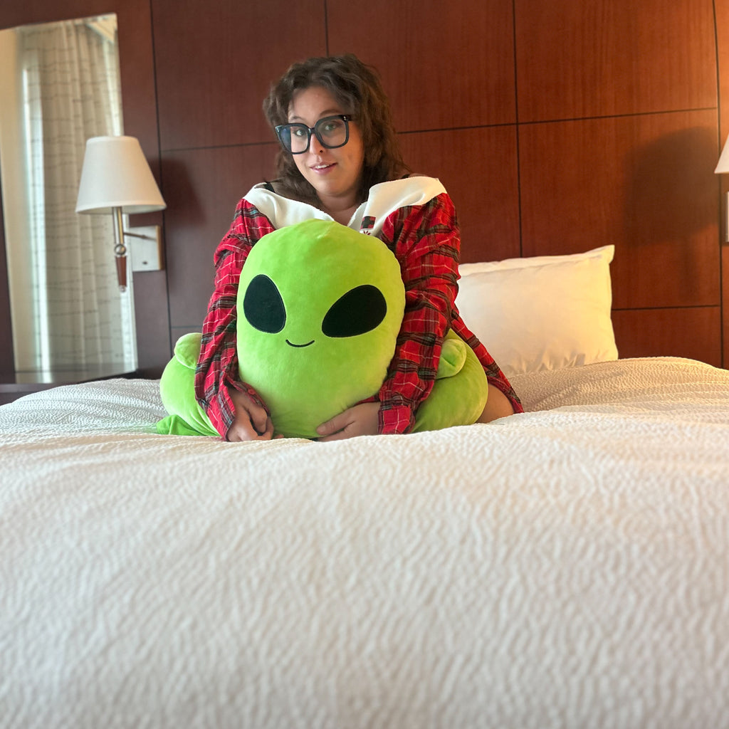 A person sitting on a bed in a hotel room holding a large green alien plush toy. The person is wearing a red and white plaid flannel shirt, and the bed has white textured bedding with a dark wood headboard in the background.