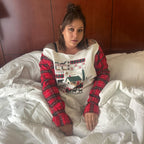 A woman sitting on a bed wearing a holiday-themed long-sleeve top with a red and black plaid pattern on the sleeves and a white front panel decorated with a Christmas village scene.
