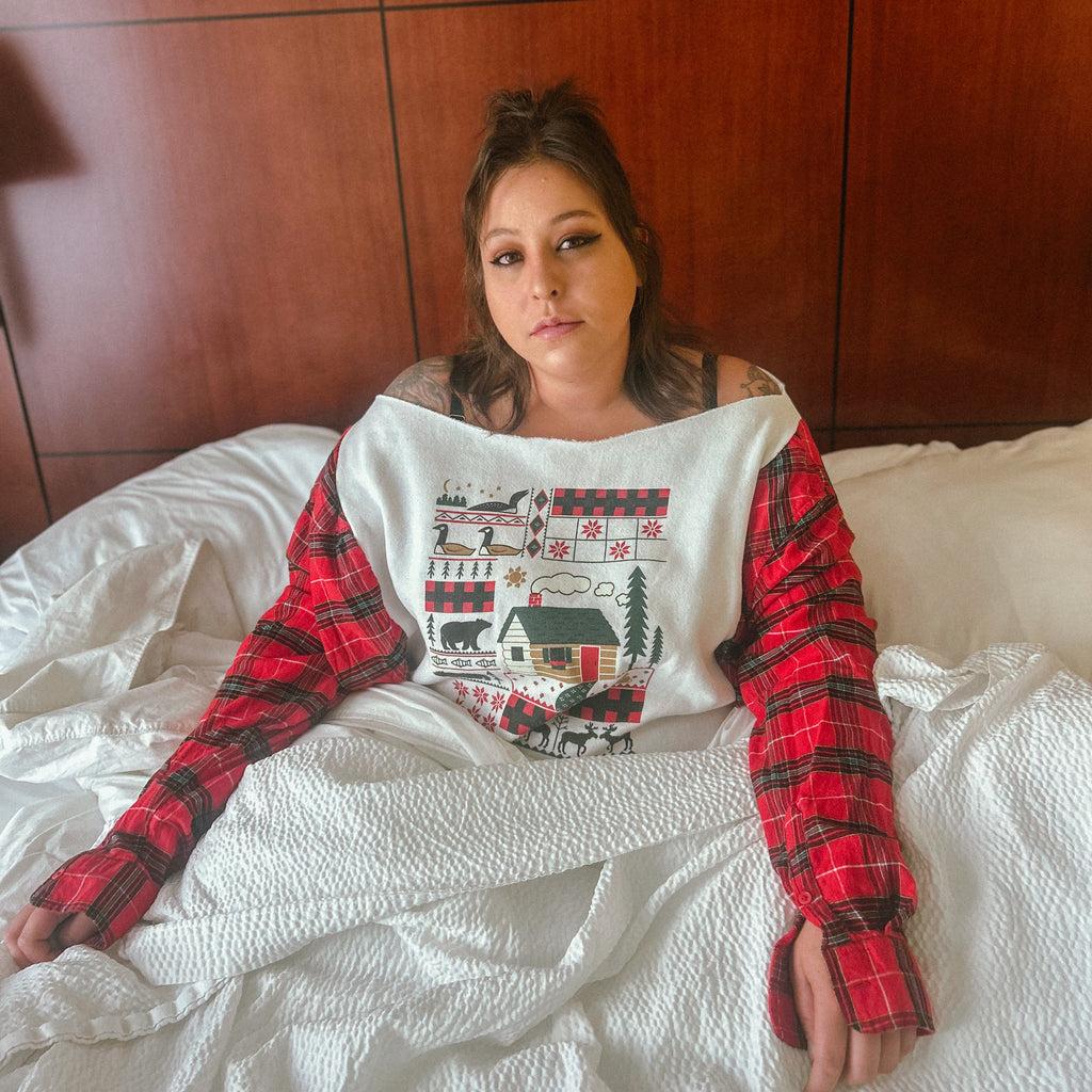 A woman sitting on a bed wearing a red and black plaid long-sleeve shirt layered under a white bib-style top with a holiday cabin graphic. The background shows a wooden headboard and white bedding.