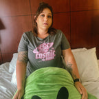 A woman sitting on a bed wearing a gray t-shirt with a pink 'Cosmic Babe' graphic, with a green plush blanket covering the foreground.