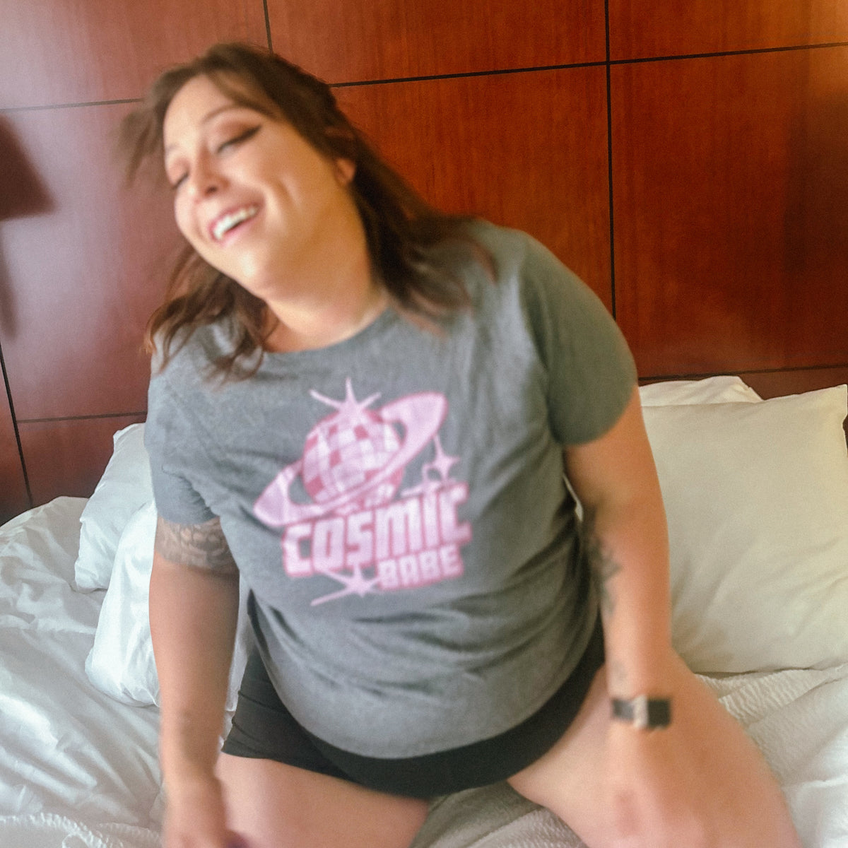 A woman smiling while wearing a gray graphic t-shirt with a pink 'Cosmic Babe' design, sitting on a bed with white sheets and a wooden headboard in the background.