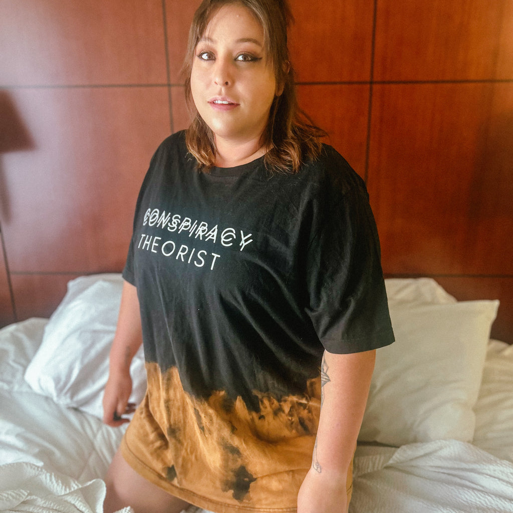 A woman wearing a black graphic t-shirt with the words 'CONSPIRACY THEORIST' in white, featuring a brown and black tie-dye gradient at the hem, sitting on a bed with white sheets.