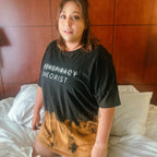 A woman wearing a black graphic t-shirt with the words 'CONSPIRACY THEORIST' in white, featuring a brown and black tie-dye gradient at the hem, sitting on a bed with white sheets.