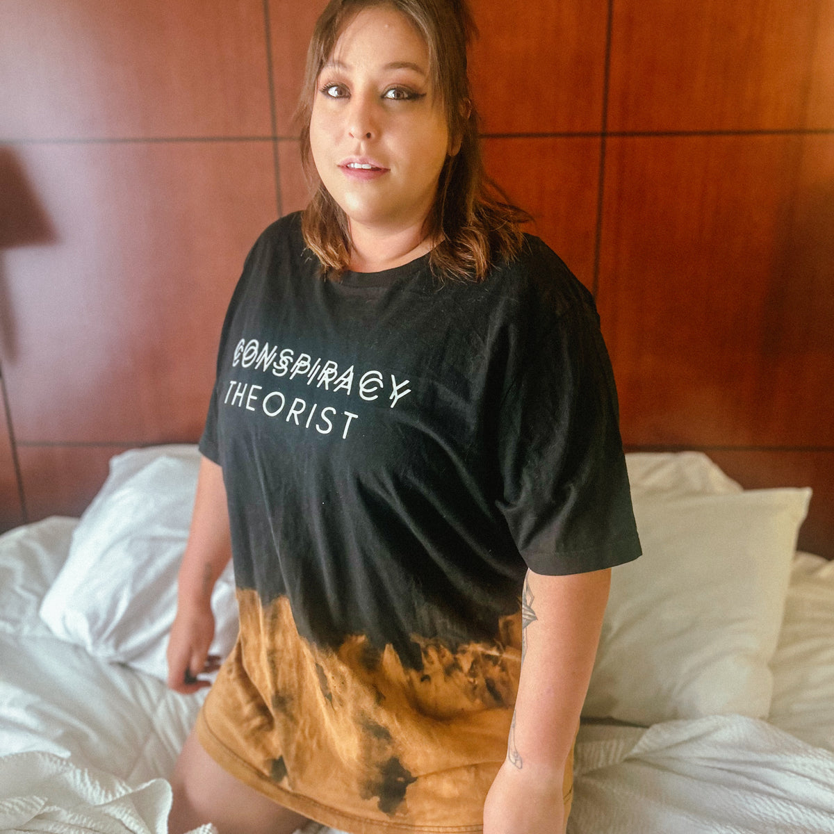 A woman wearing a black graphic t-shirt with the words 'CONSPIRACY THEORIST' in white, featuring a brown and black tie-dye gradient at the hem, sitting on a bed with white sheets.