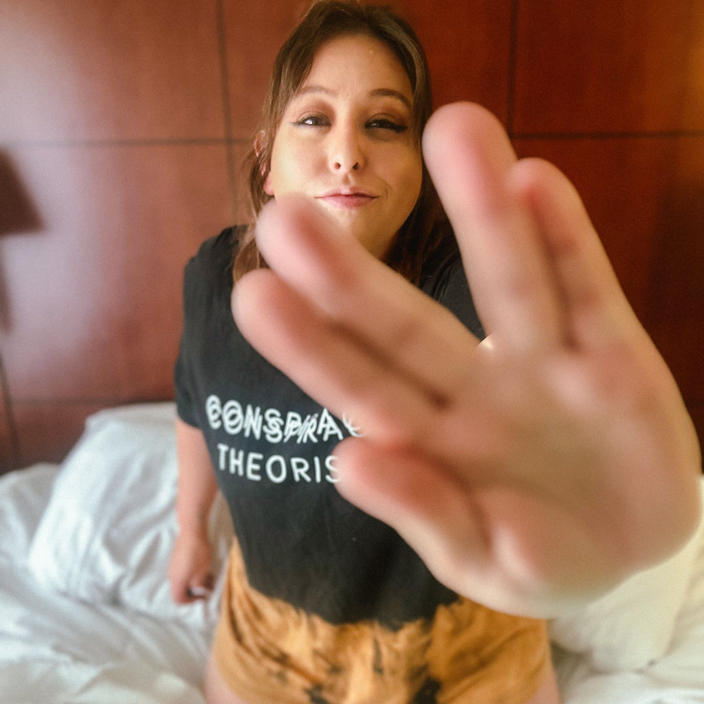 A woman wearing a black graphic t-shirt with the text 'BONSAI THEORIS' and orange tie-dye shorts, sitting on a bed with white sheets and a wooden headboard in the background, with her hand reaching towards the camera.