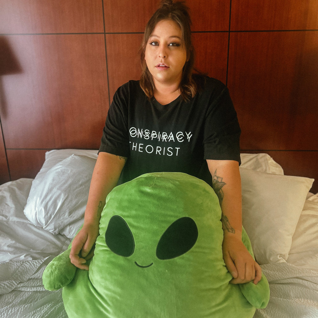 A woman sitting on a bed in a bedroom, holding a large green alien plush toy. She is wearing a black t-shirt with the text '@SPIRITAY THEORIST' printed in white.