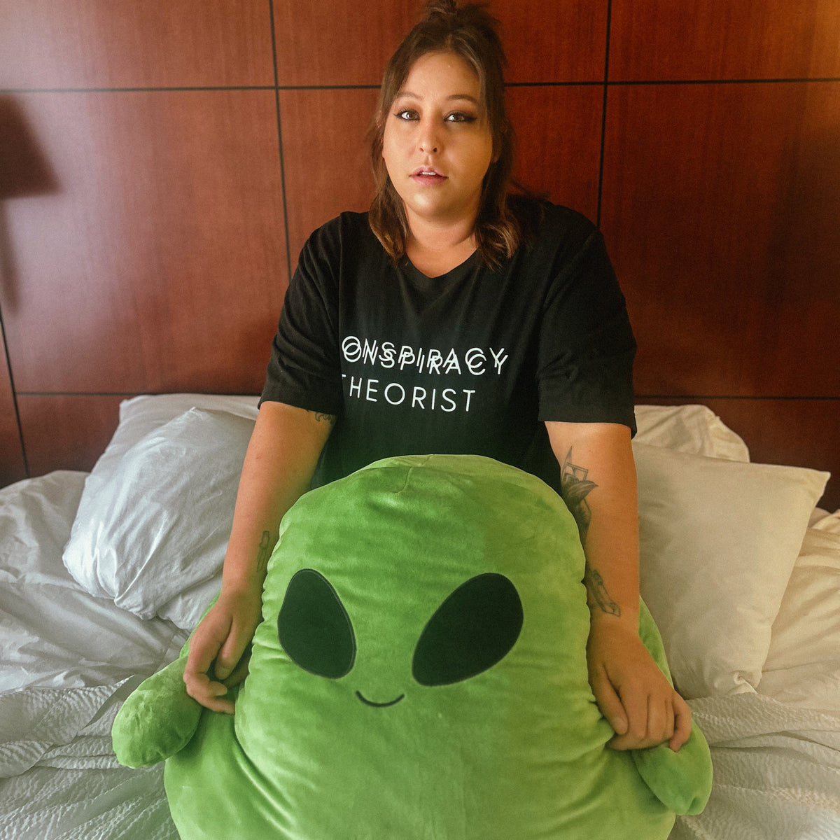 A woman sitting on a bed in a bedroom, holding a large green alien plush toy. She is wearing a black t-shirt with the text '@SPIRITAY THEORIST' printed in white.