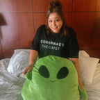 A woman wearing a black t-shirt with the text 'CONSPIRACY THEORIST' sits on a bed holding a large green alien plush pillow.