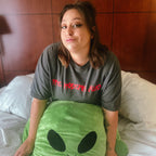A woman sitting on a bed wearing a dark gray t-shirt with red text that reads 'THE MODERN ALIEN' and holding a large green alien plush toy.