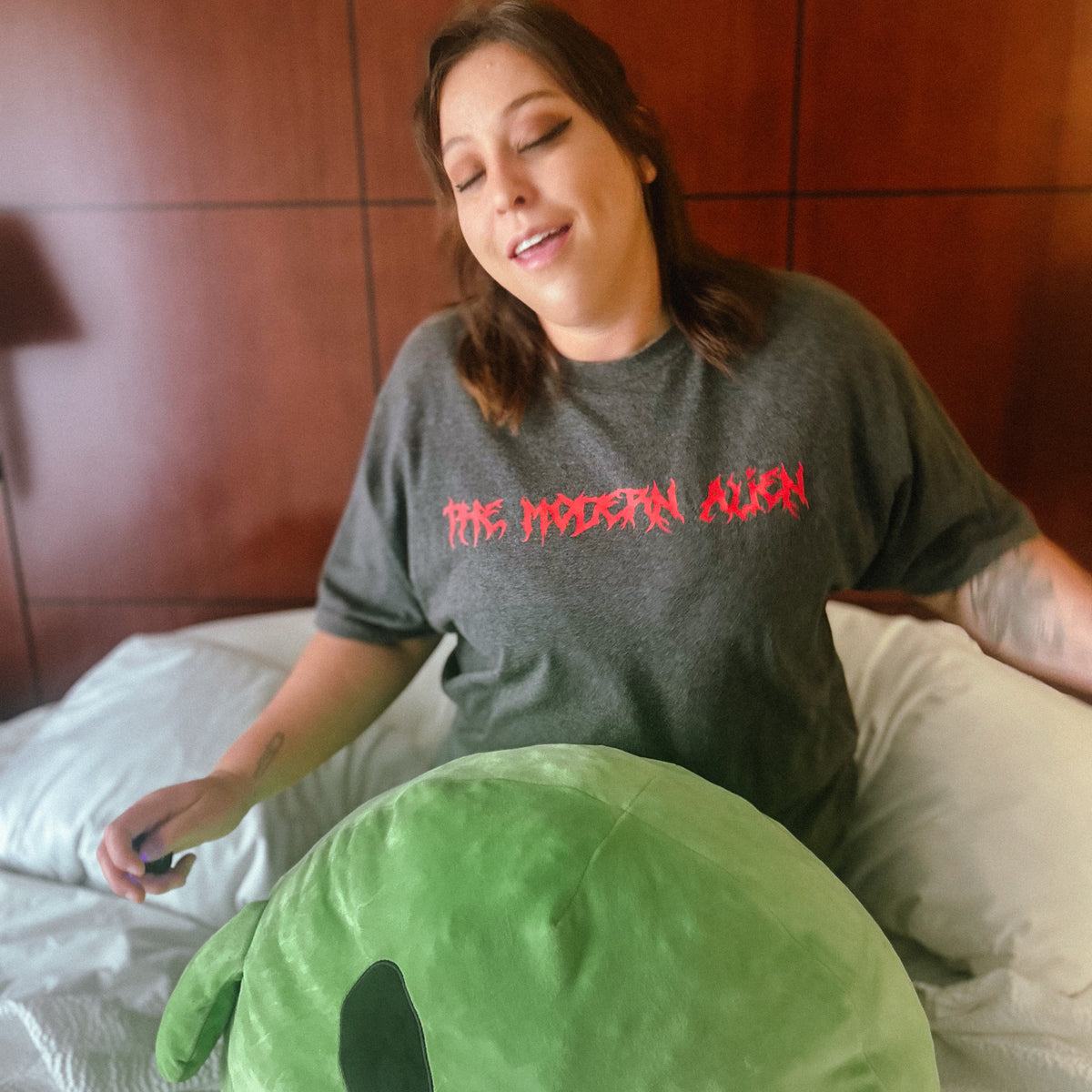 A woman sitting on a bed wearing a dark gray t-shirt with 'THE MODERN ALIEN' printed in red across the chest, holding a large green alien plush toy.