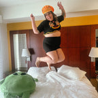 A woman jumps excitedly on a bed in a hotel room, wearing a black graphic crop top, black shorts, and an orange patterned hat. A large green plush toy sits on the white bedding.