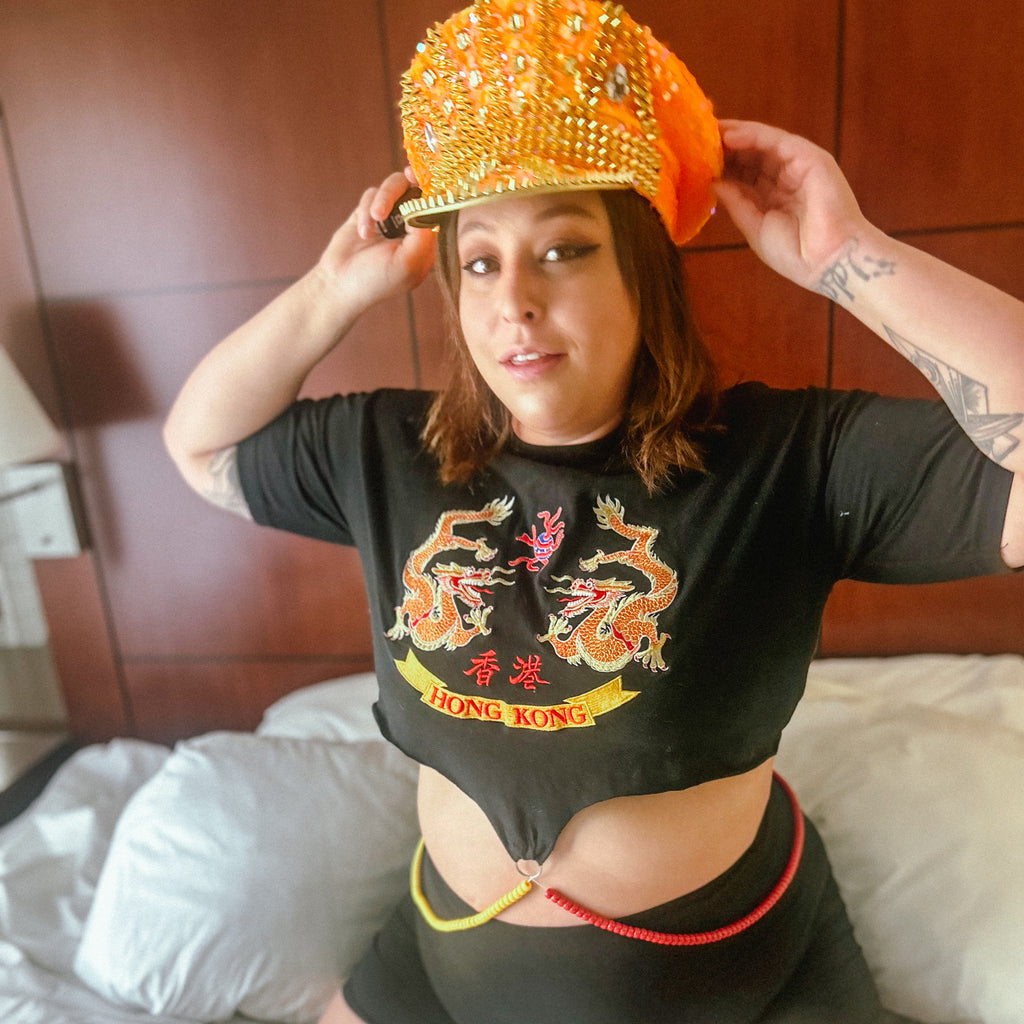 A woman wearing a black crop top with a colorful embroidered dragon graphic and 'HONG KONG' text, paired with black shorts and an orange embellished hat, sitting on a bed.