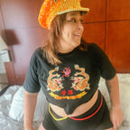 A woman wearing a black cropped t-shirt with a gold and red dragon graphic and Chinese characters, paired with a black skirt featuring red and yellow trim, and a gold beaded hat, sitting on a bed.